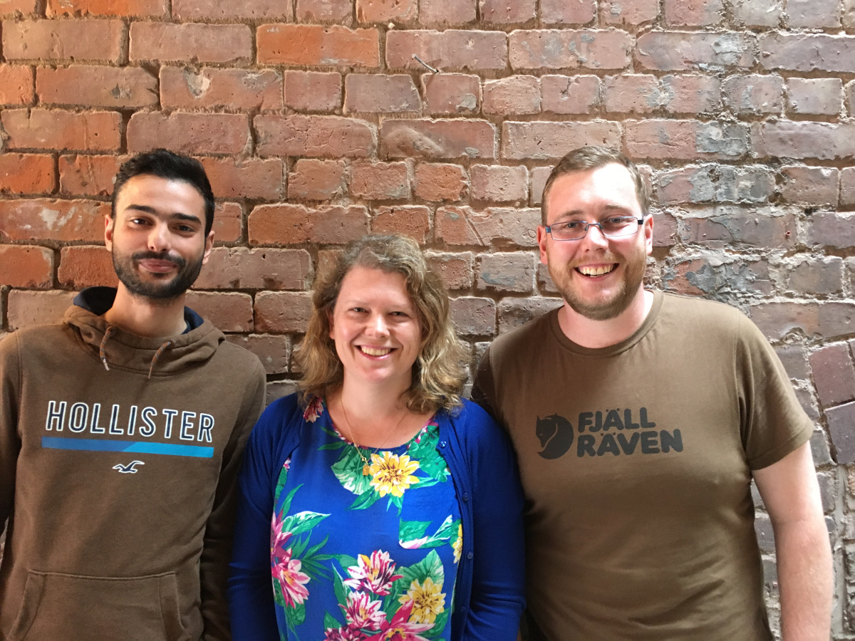 Three adults, Georgios, Sophie, Tom, staring into the camera smiling stood infront of a brick wall