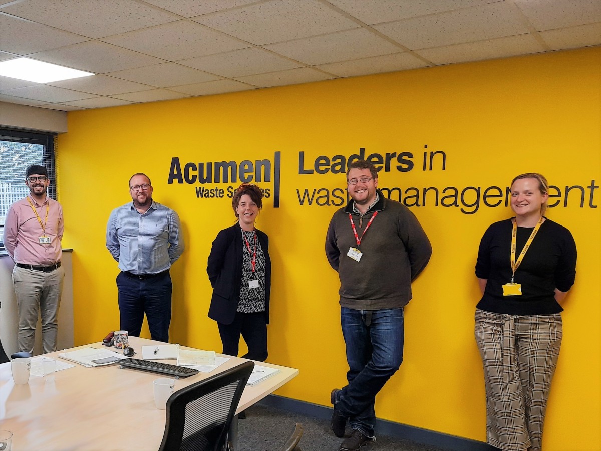 Photograph of business people smiling at camera Michael Ruddock, Kris Sutton, Alexa Culver, Tom Passmore, Lauren Hill in front of yellow wall with Acumen Waste Services Leaders in waste management written